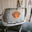 A gray hard hat with an orange and black monkey face logo rests on equipment in a workshop, featuring the El Mono Hook Pad secured with industrial-strength adhesive, as tools hang on a wooden wall in the background.