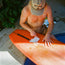 A shirtless person wearing a beige HERMANOS hat sits near a pool, waxing an orange surfboard decorated with Howler Brothers stickers. Lush green plants are nearby, and sunlight casts shadows on the ground.