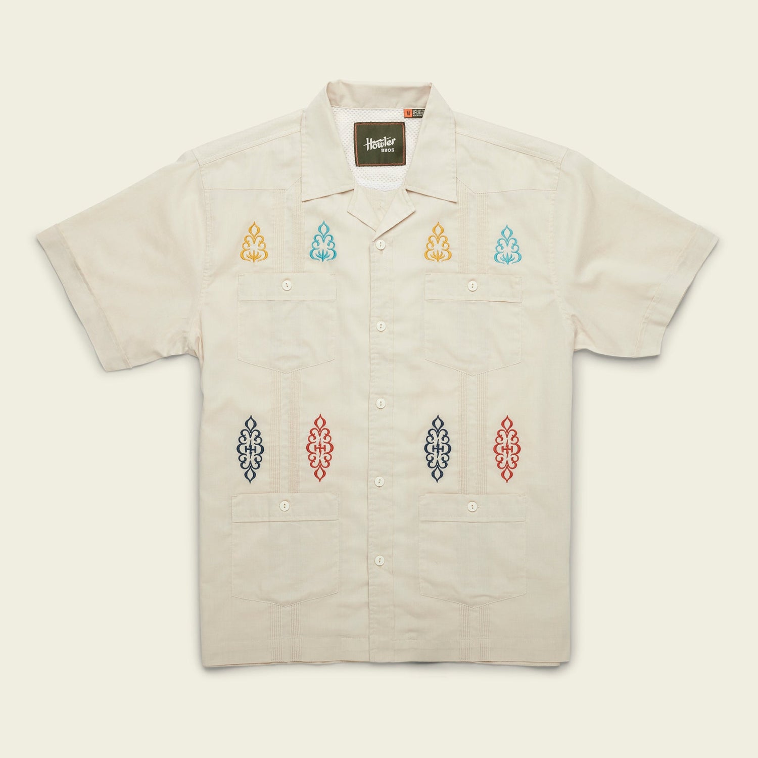 A modern fit, short-sleeve Guayabera shirt in light beige, featuring four front pockets and colorful embroidered patterns on the chest and lower front, crafted from technical cotton-poly stretch fabric for added comfort.