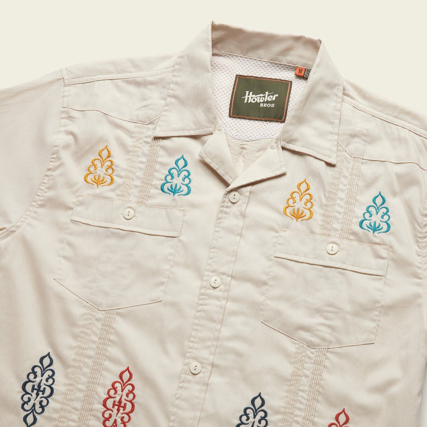 A light beige short-sleeve shirt with a modern fit, two chest pockets, pointed collar, and colorful embroidered patterns in blue, orange, red, and navy. Crafted from technical cotton-poly stretch fabric. The label inside reads Howler Bros.