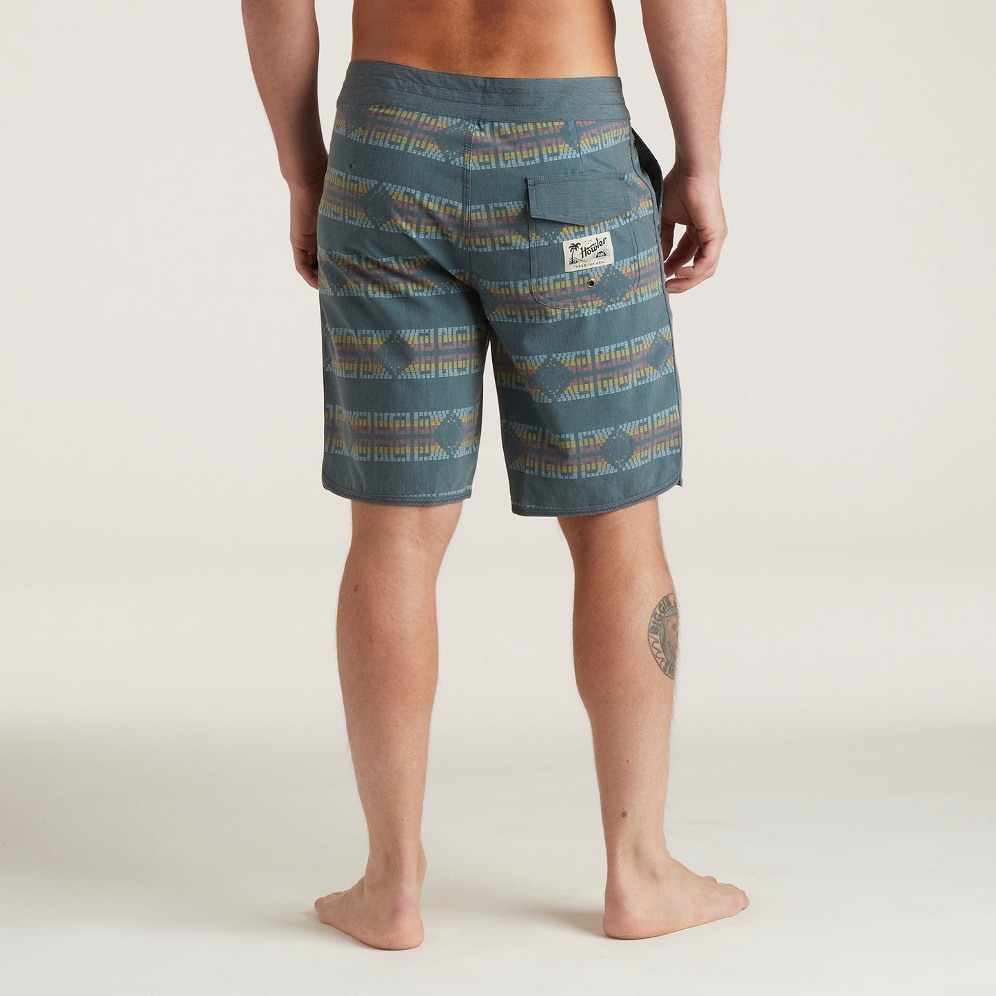 A person is standing barefoot on a light surface, shown from the back, wearing Bruja Boardshorts—patterned blue-green knee-length shorts with a back pocket and visible leg tattoo. The background is plain and light-colored.
