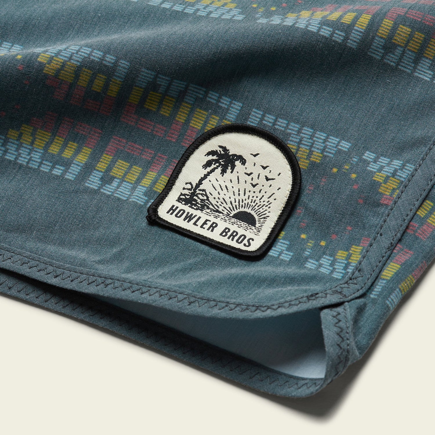 Close-up of blue fabric from the Bruja Boardshorts, featuring a colorful geometric pattern and a sewn patch with a palm tree, sun rays, and the text “HOWLER BROS.”.