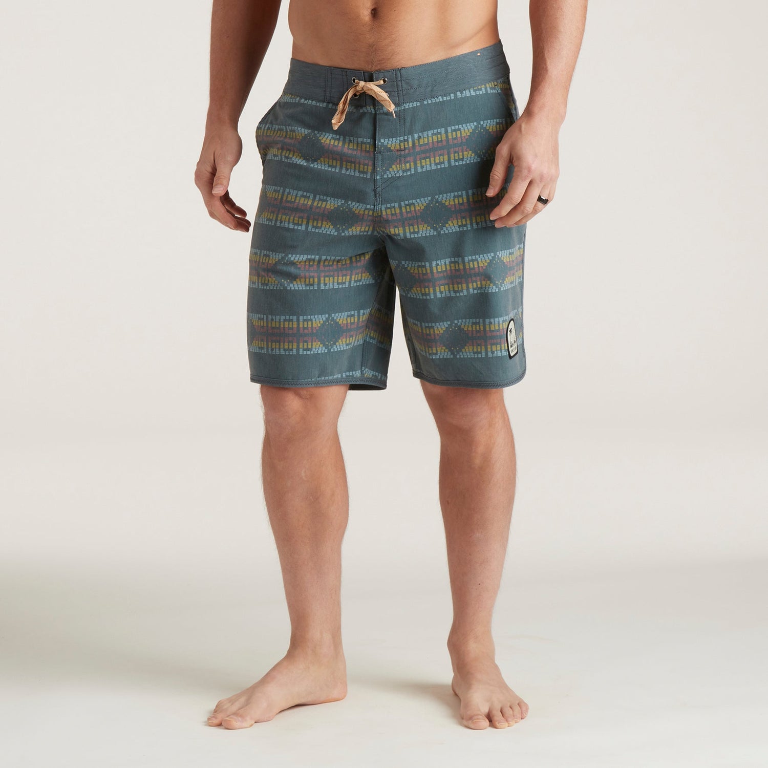 A man stands barefoot in blue patterned Bruja Boardshorts with a drawstring waist and a patch on the left leg, against a plain light background. His torso is unclothed and his arms are relaxed at his sides.