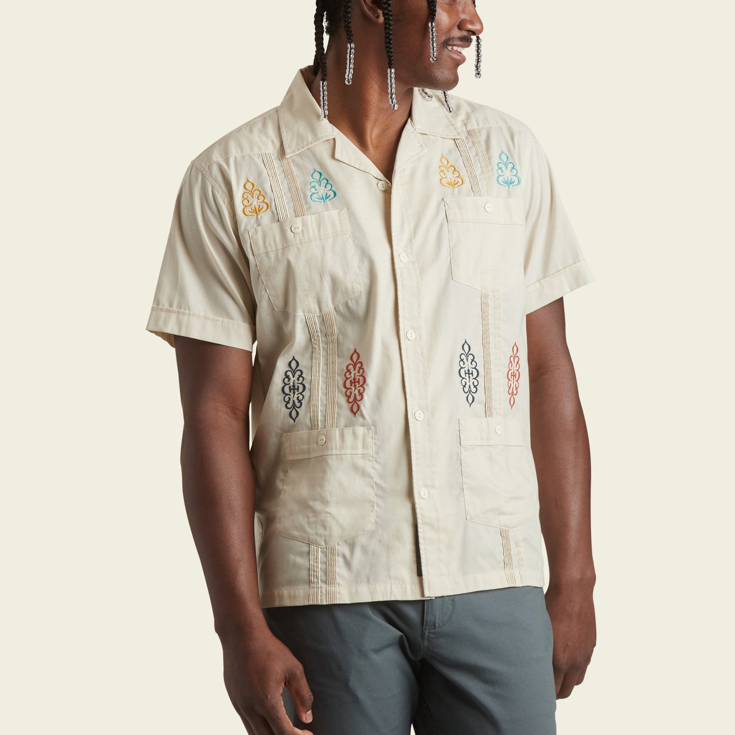 A person wearing a modern fit, short-sleeve cream-colored Guayabera with colorful embroidered patterns, standing with one arm relaxed and looking to the side.