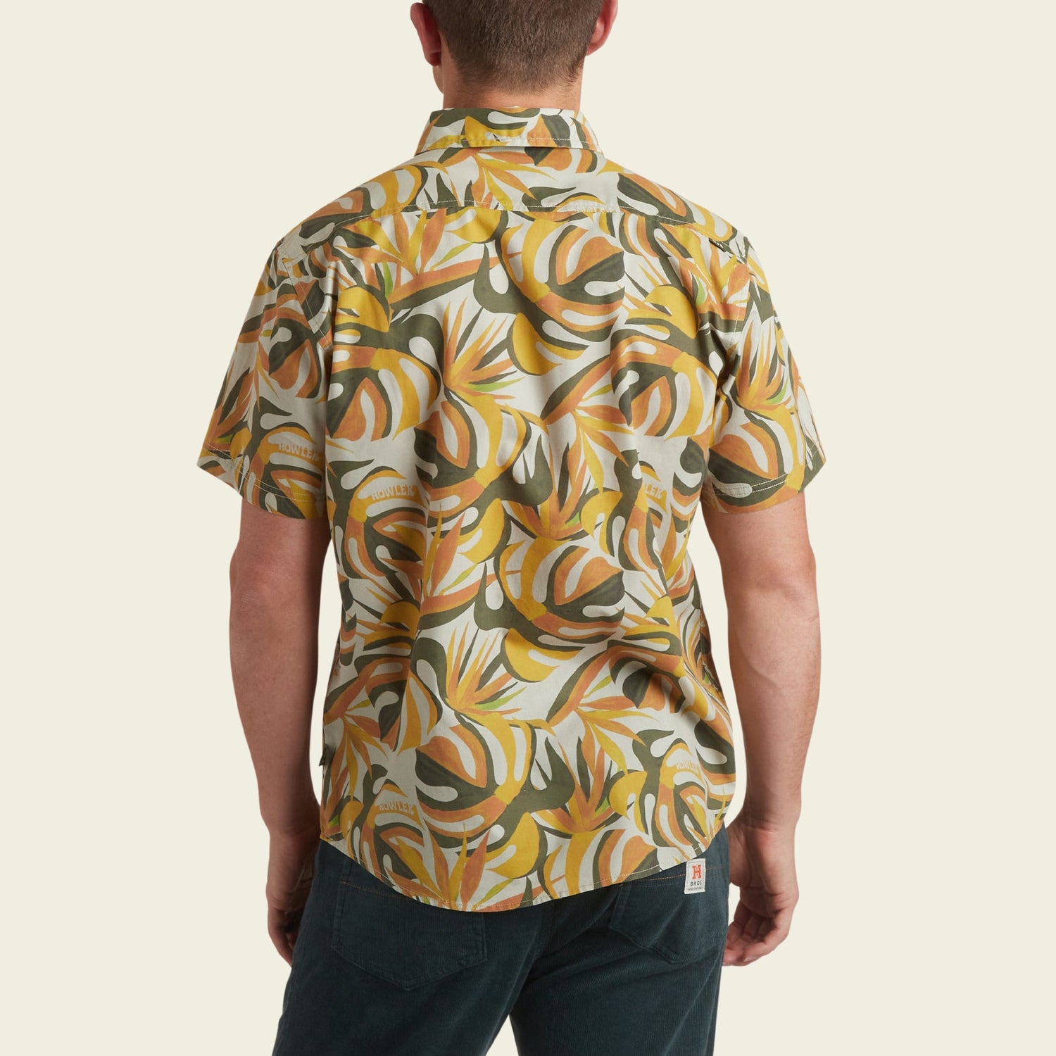 A man wearing the Mansfield Shirt, a lightweight cotton-poly shirt with a bold yellow, orange, and green abstract pattern, is viewed from the back against a plain light background—perfect for showcasing unique seasonal patterns.