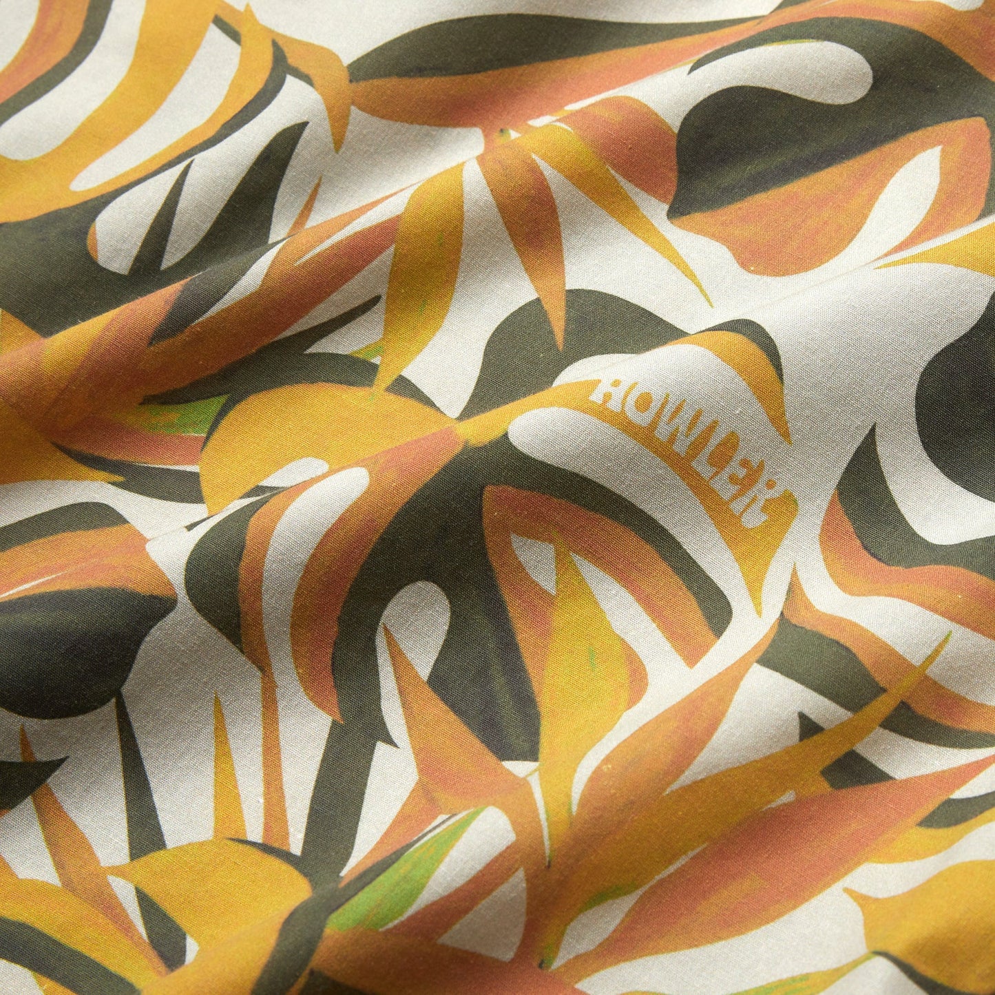 A close-up of fabric with a tropical leaf pattern in yellow, orange, and green tones. The word HOWLER is partially visible among the leaves of this lightweight cotton-poly shirt, featuring unique seasonal patterns. The material appears slightly wrinkled.