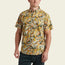A man wearing the Mansfield Shirt—a short-sleeve button-up with yellow, green, and white tropical leaf print—stands against a plain light background. This lightweight cotton-poly shirt showcases unique seasonal patterns perfect for warm weather.