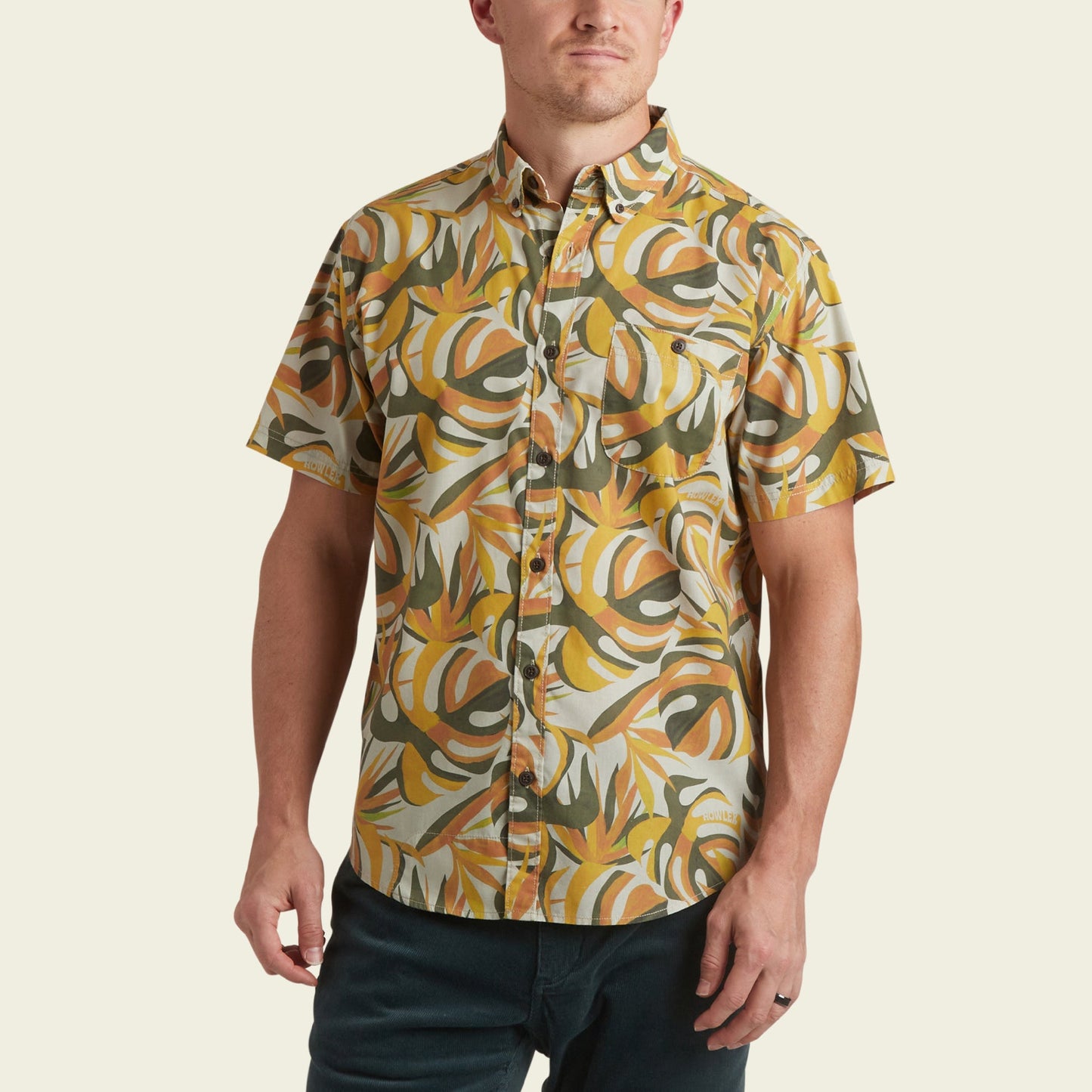 A man wearing the Mansfield Shirt—a short-sleeve button-up with yellow, green, and white tropical leaf print—stands against a plain light background. This lightweight cotton-poly shirt showcases unique seasonal patterns perfect for warm weather.