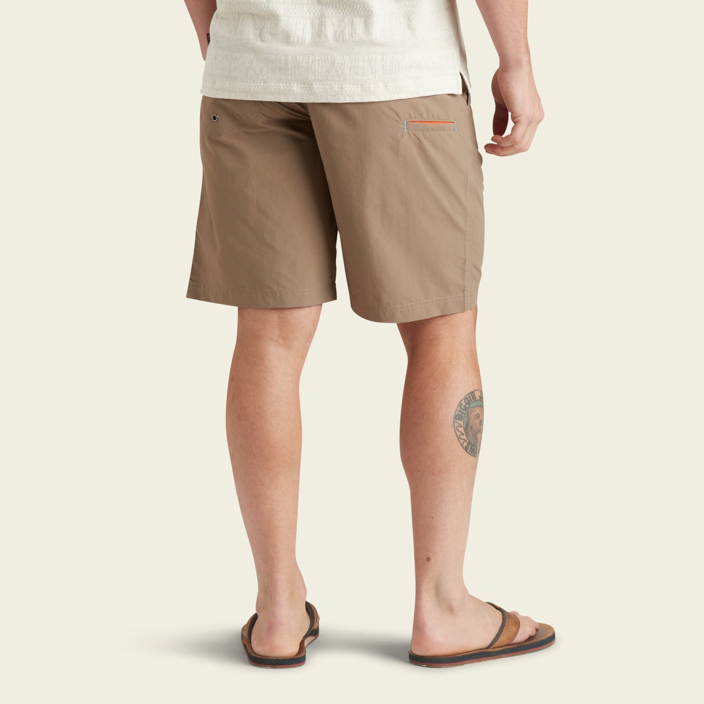 A person stands facing away, wearing Horizon Hybrid Shorts in light brown—versatile shorts crafted from ripstop fabric. The shorts have a back pocket with a small accent. A circular tattoo is visible on their left calf against an off-white background.
