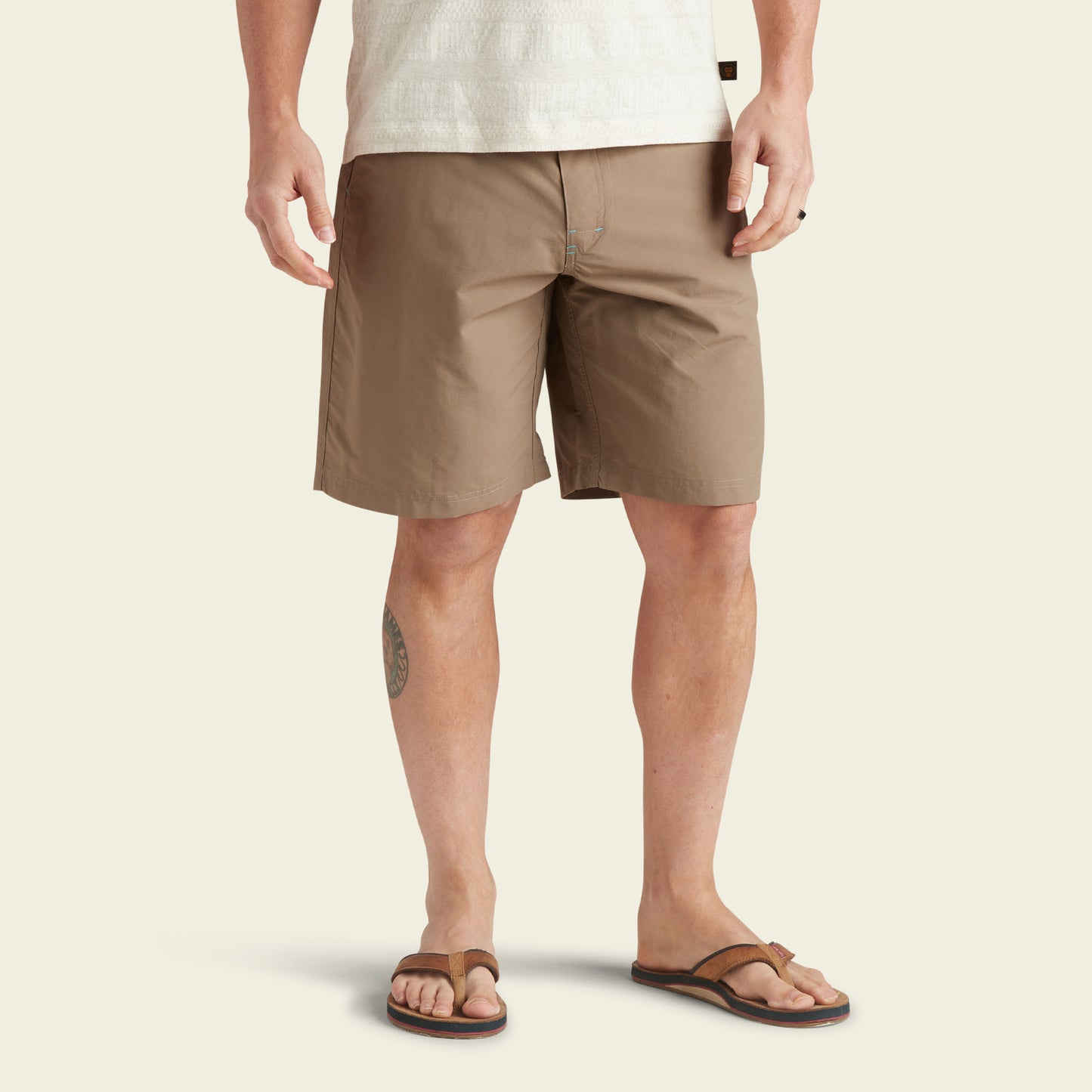 A person wearing a white textured shirt, versatile Horizon Hybrid Shorts made from ripstop fabric, and brown flip-flops stands against a plain light background. Only the lower half of their body is visible.