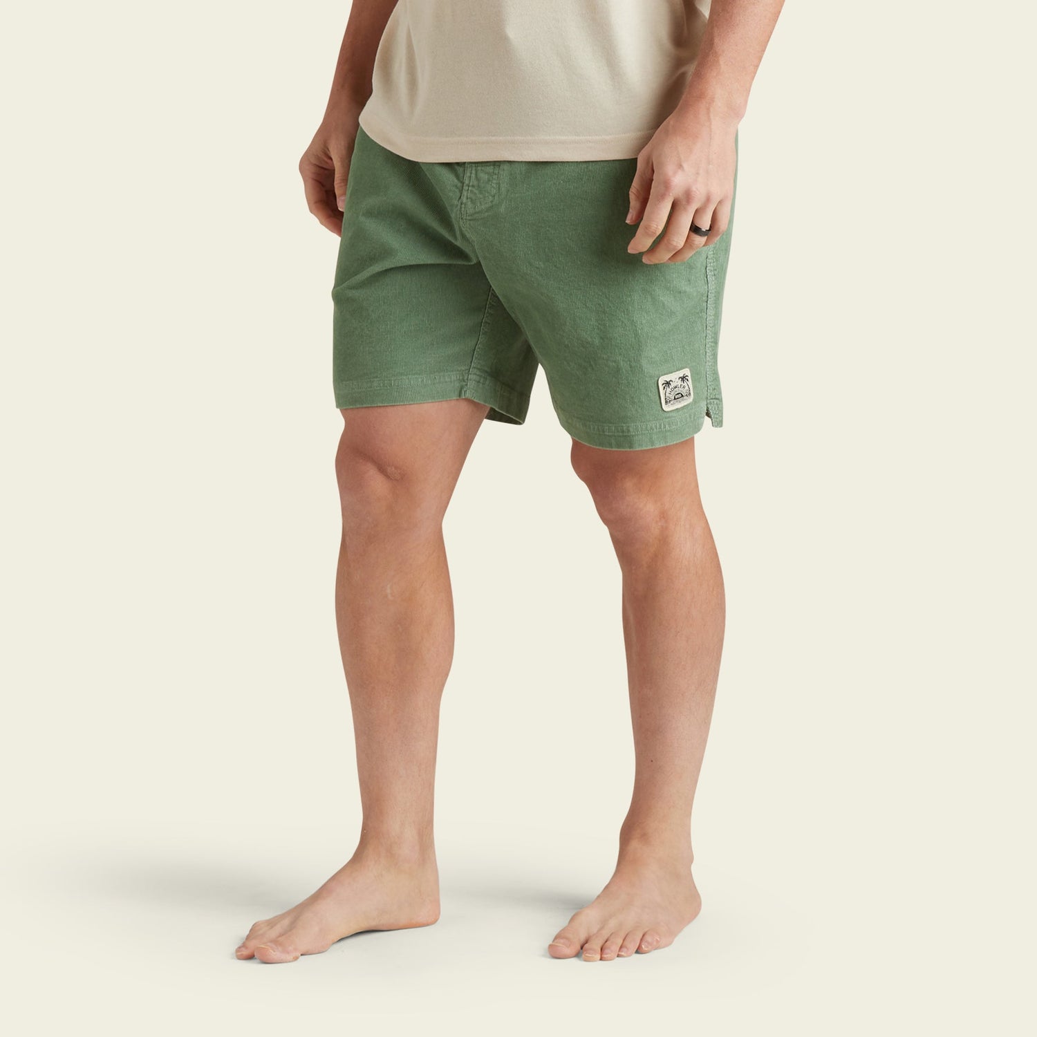 A person stands barefoot, wearing Pressure Drop Cord Shorts in green—crafted from stretch cotton corduroy for comfortable wear—and a beige t-shirt. The shorts feature a small white patch on the left leg. The background is plain and light-colored.