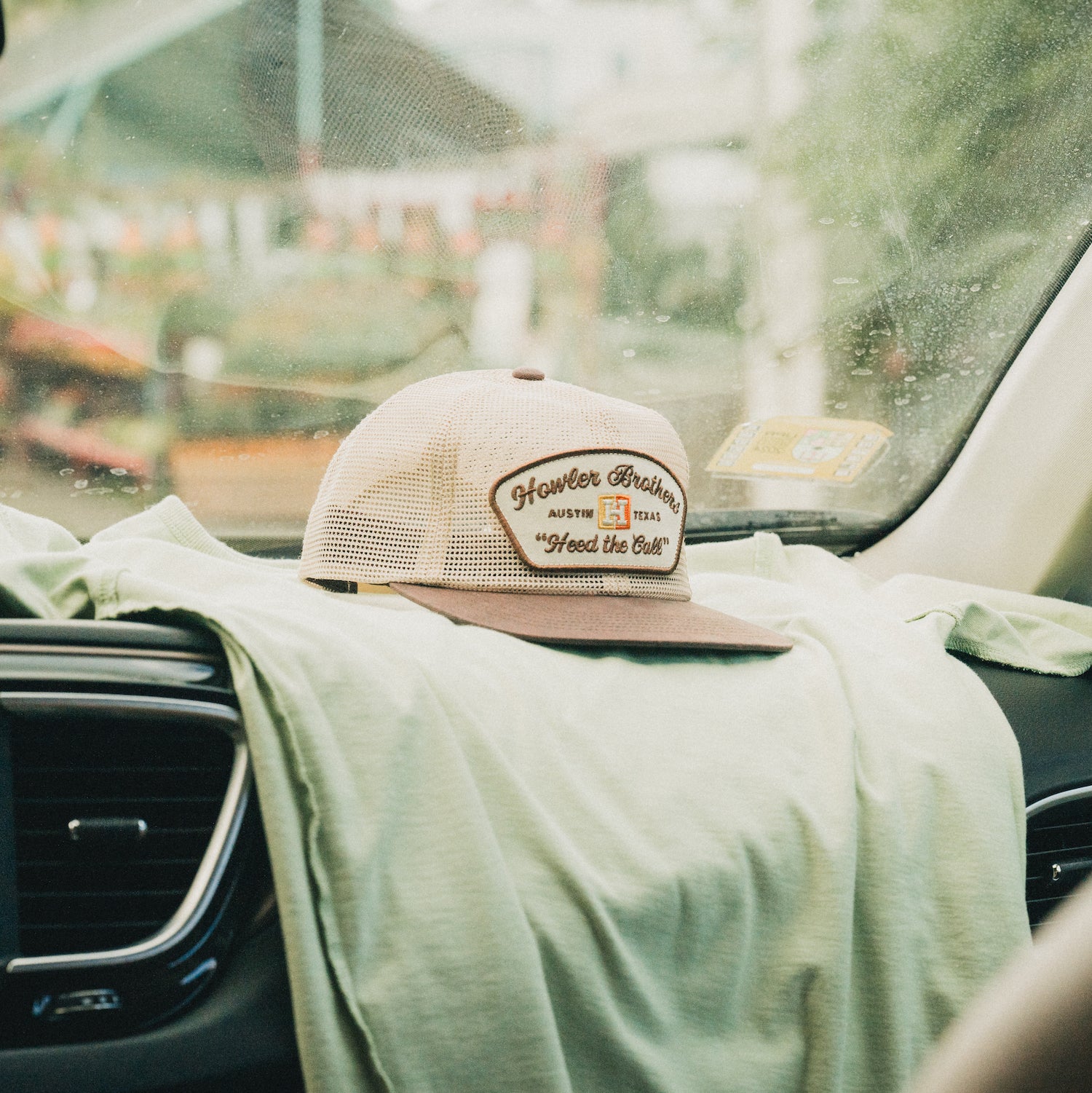 A beige and brown Howler Brothers trucker hat with an unstructured snapback and soft crown rests on a light green shirt draped over a car dashboard. The adjustable snapback closure shows in the cozy, blurred backdrop of greenery outside.