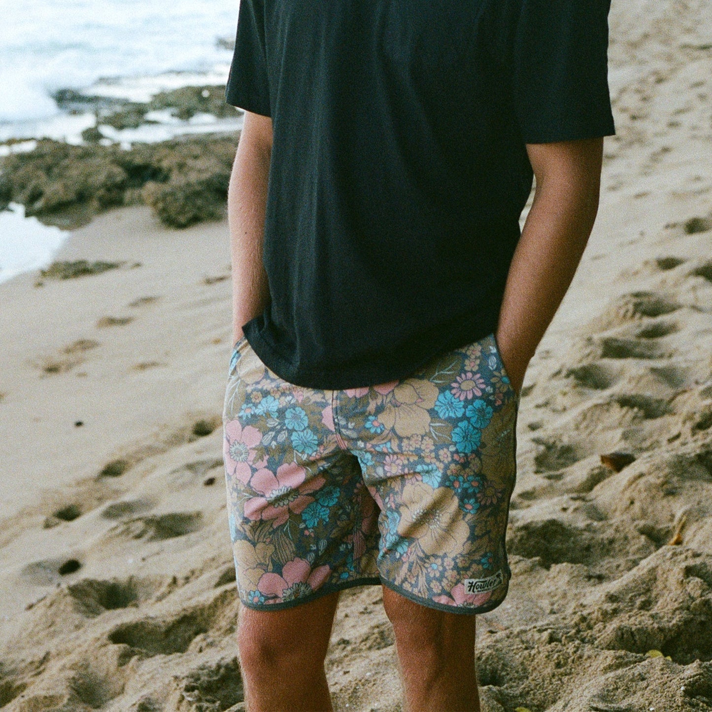A person stands on a sandy beach with hands in the draining front pockets of floral-patterned Bruja Boardshorts, wearing a plain black T-shirt. The ocean and some rocks are visible in the background.