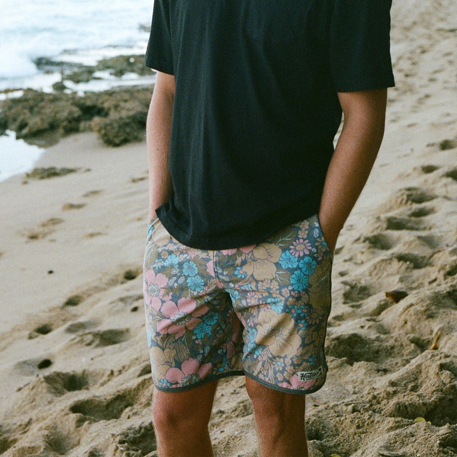 A person stands on a sandy beach with hands in the draining front pockets of floral-patterned Bruja Boardshorts, wearing a plain black T-shirt. The ocean and some rocks are visible in the background.