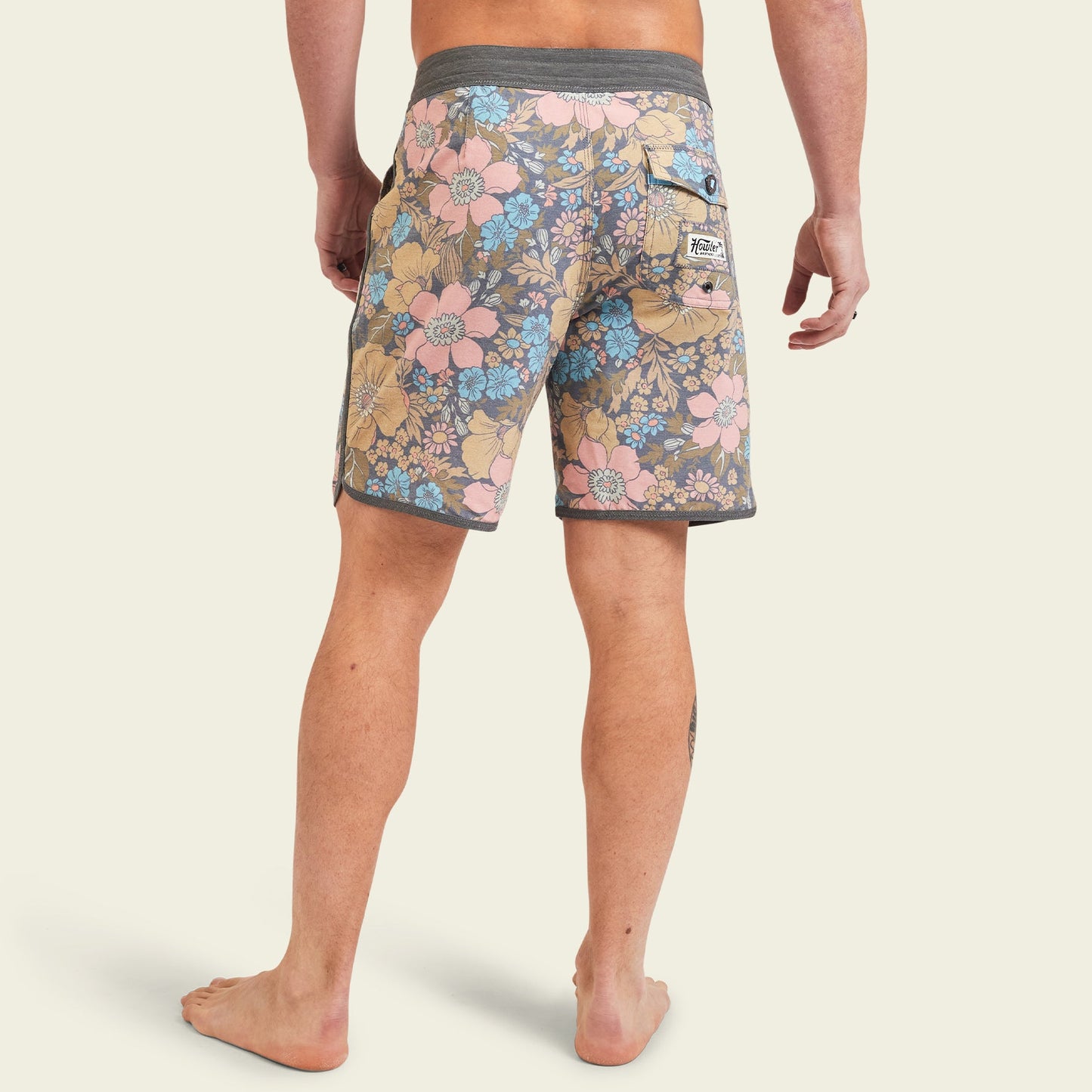 A person stands barefoot, facing away, wearing Bruja Boardshorts in pink, blue, and tan floral print with a gray waistband and trim. Made from stretchy fabric, these shorts feature draining front pockets and a back pocket with a white label.