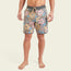 A man stands barefoot in Bruja Boardshorts, made from stretchy fabric with draining front pockets, featuring floral patterns in pink, yellow, blue, and brown with a gray waistband and drawstring against a plain light background.