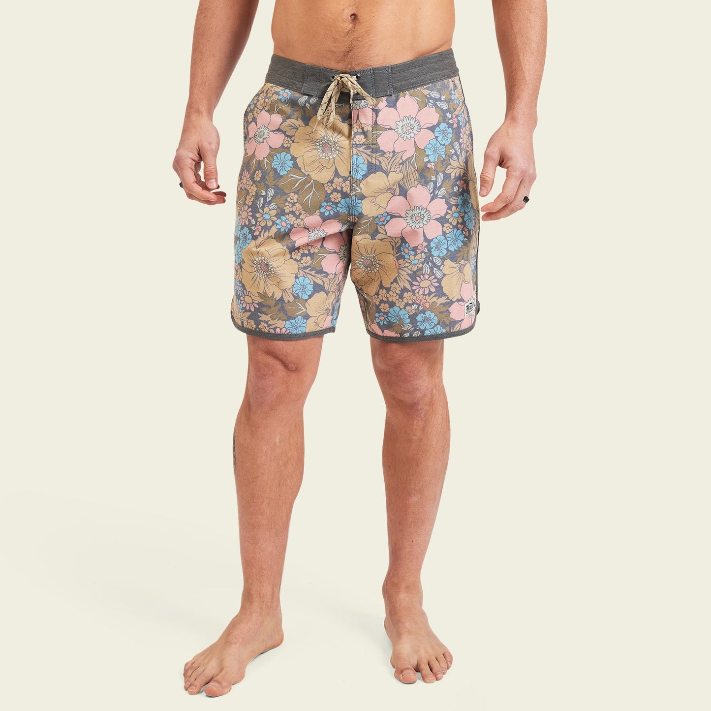 A man stands barefoot in Bruja Boardshorts, made from stretchy fabric with draining front pockets, featuring floral patterns in pink, yellow, blue, and brown with a gray waistband and drawstring against a plain light background.