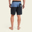 A barefoot person stands facing away, wearing black and blue color-blocked Buchannon Boardshorts with a back pocket and a small patch logo above the pocket, perfect quick drying surf shorts against a plain light background.