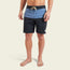 A person wearing Buchannon Boardshorts—the quick drying, two-tone surf shorts with a blue upper section, black lower section, drawstring waist, and a small patch near the left hem—stands barefoot against a plain background.