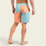 A person stands barefoot, showing the back of their legs and wearing quick drying boardshorts in peach and light blue with a pocket and logo on the right side. A tattoo is visible on the back of their left calf.