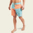 A man stands barefoot wearing quick drying surf shorts in light blue and orange with a drawstring waist and a patch label near the left hem. The background is plain and light-colored.