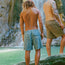 Two people in swim trunks with elastic waistbands stand with their backs to the camera at the edge of a pool beneath a waterfall. One person has curly hair and wears a hat; both are looking at the water.