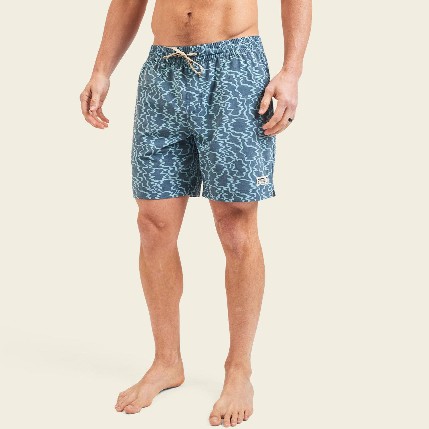A person stands barefoot wearing blue patterned beach shorts with an elastic waistband and drawstring, set against a plain light background.