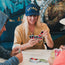 Three people sit around a table playing Uno. The person in the center, wearing a vintage-inspired high crown hat and tan shirt, smiles while holding their cards. Two others, partially visible, also hold Uno cards. A colorful mural is in the background.
