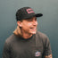 A man with long blond hair, a mustache, and an adjustable snapback hat with a glare-cutting brim smiles while looking to the side. He wears a dark t-shirt with a small colorful Howler artwork logo on the pocket, standing against a teal wall.