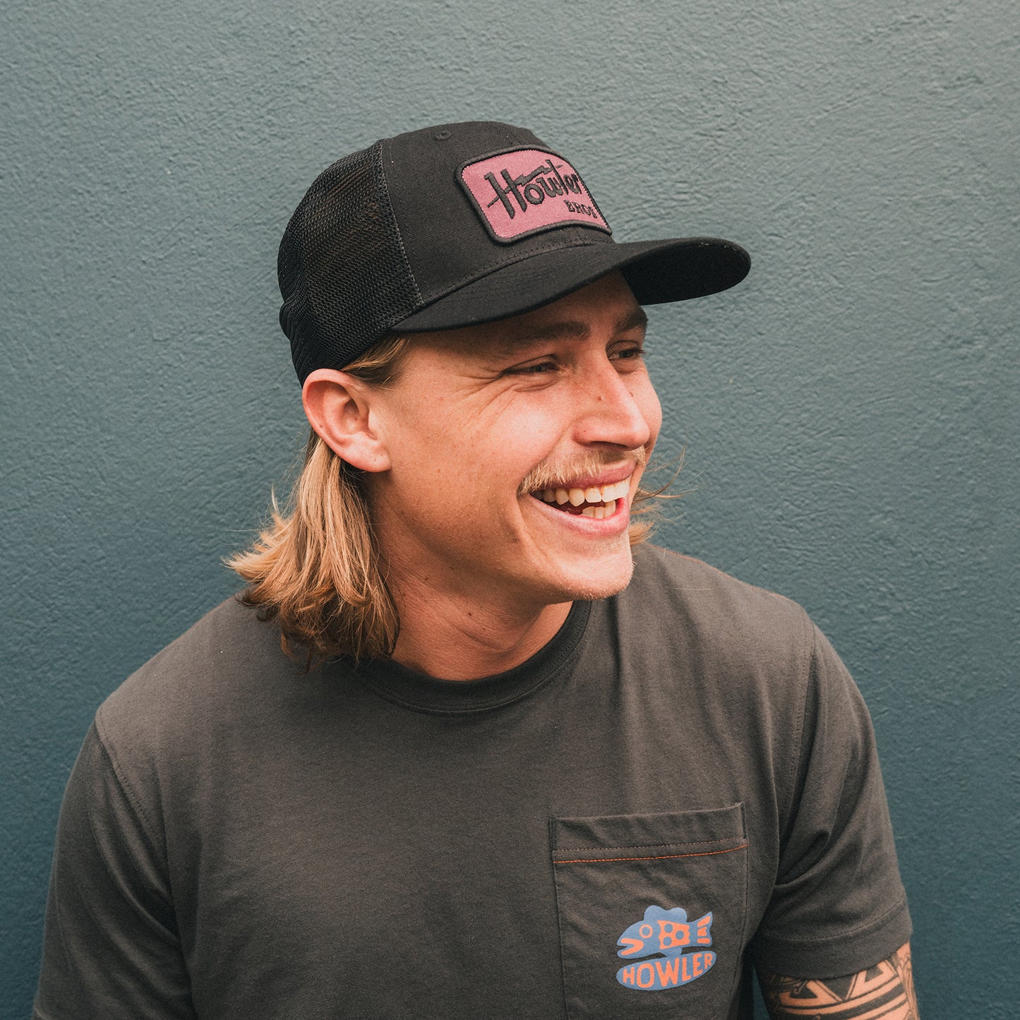 A man with long blond hair, a mustache, and an adjustable snapback hat with a glare-cutting brim smiles while looking to the side. He wears a dark t-shirt with a small colorful Howler artwork logo on the pocket, standing against a teal wall.