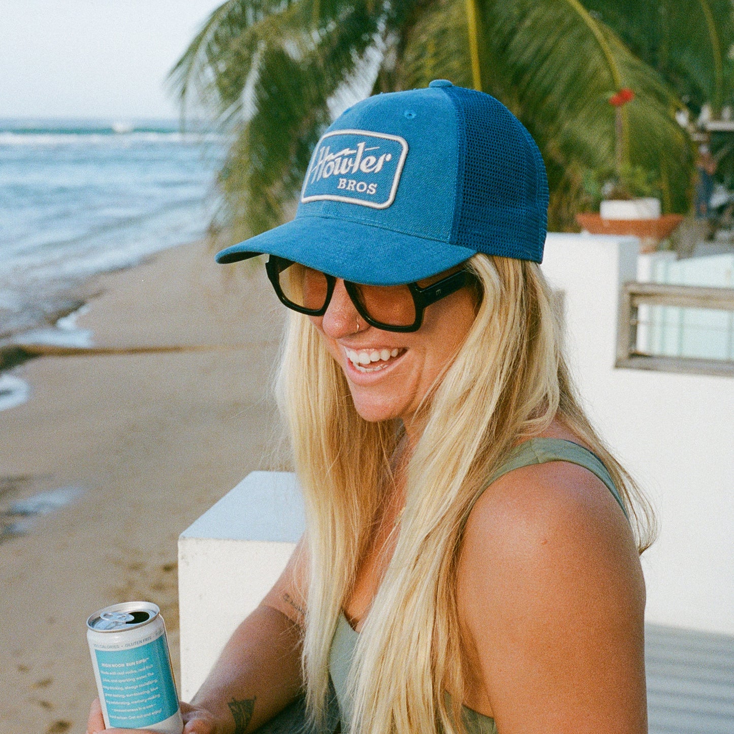 A smiling woman with long blonde hair, wearing a blue Howler Bros cap with a glare-cutting brim and sunglasses, holds a can while standing by the beach with palm trees and ocean in the background.