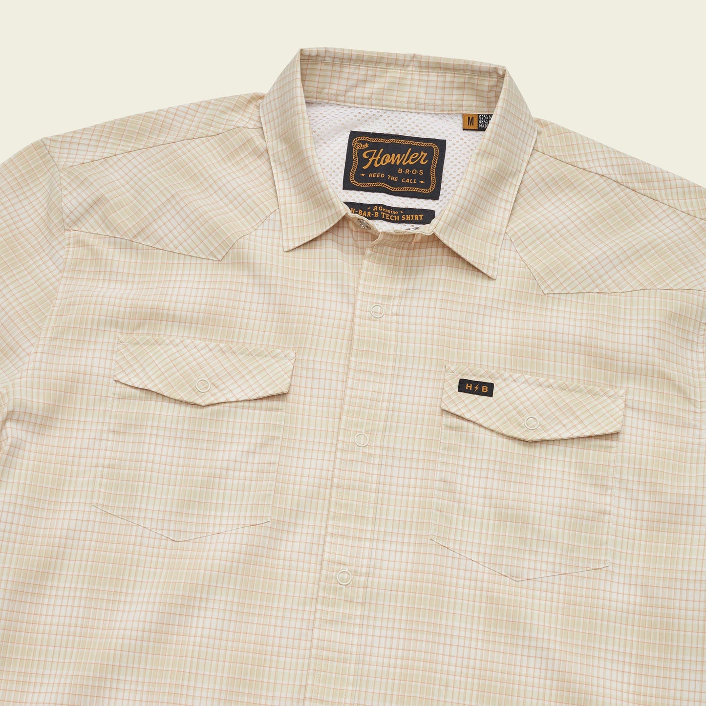 A beige plaid lightweight stretch shirt with a collar, two chest pockets with button flaps, a vented yoke for breathability, and a visible Howler Bros brand label on the inside collar.