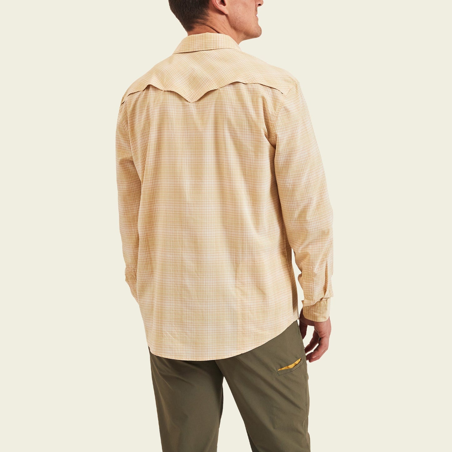 A man facing away, wearing a light beige plaid lightweight stretch shirt with a vented yoke and olive green pants, standing against a plain light background.