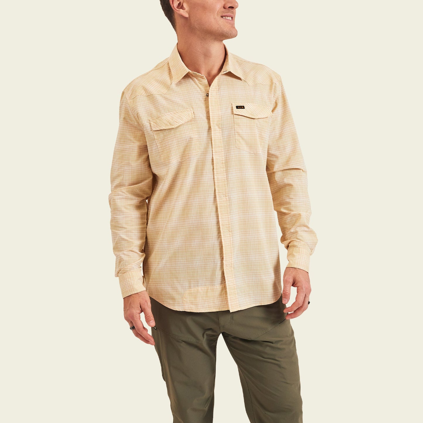 A man wearing a light yellow plaid lightweight stretch shirt with long sleeves and olive green pants stands against a plain, light background.