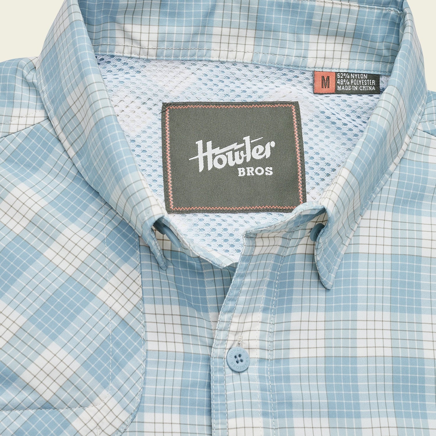 A light blue and white plaid Matagorda Shirt with a mesh inner lining, featuring a Howler Bros label and a size medium tag sewn inside the collar. This travel shirt is quick drying for comfort on the go.