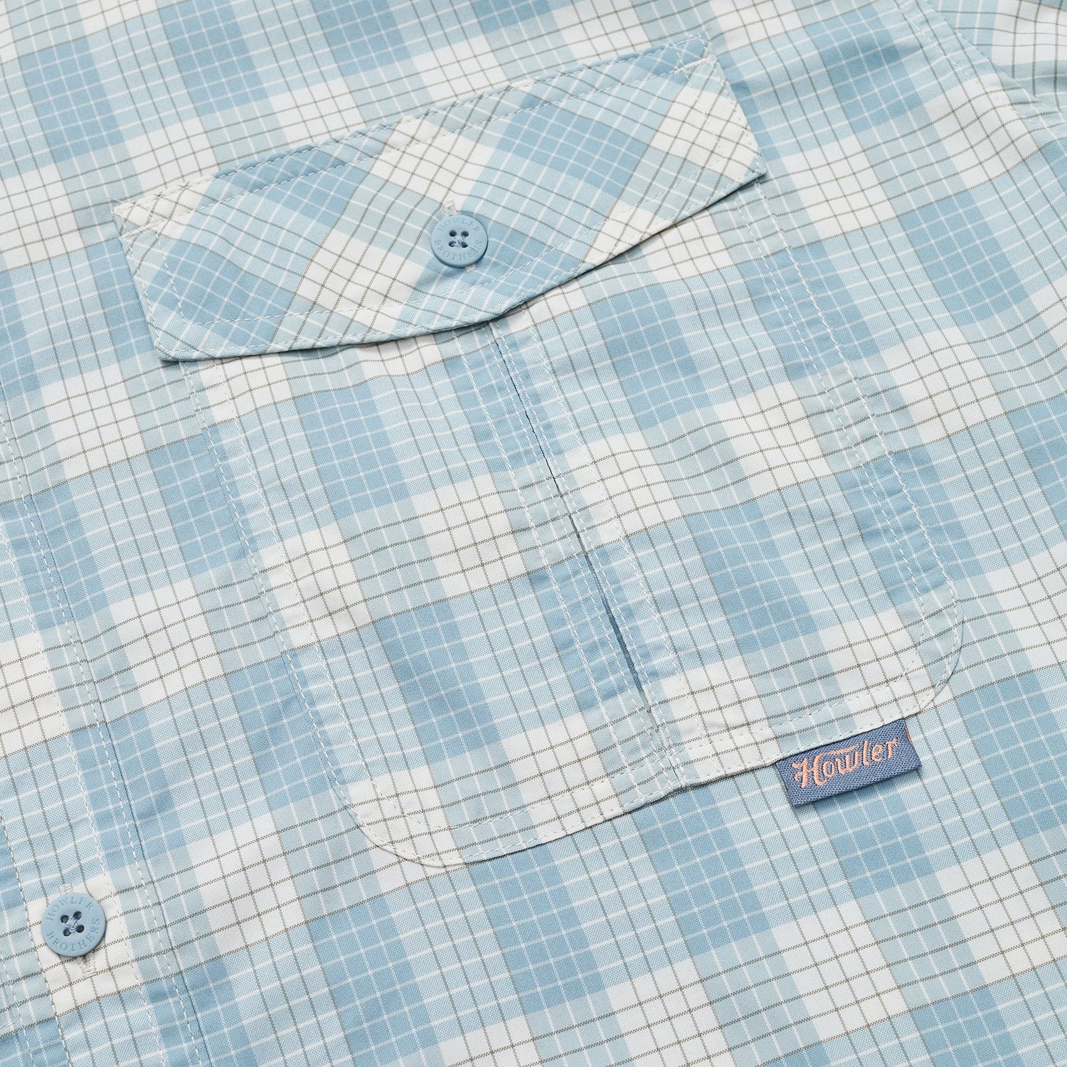 Close-up of a light blue and white checkered Matagorda Shirt with a buttoned chest pocket. The pocket features a blue button and a small fabric tag with the word Howler stitched in red. Ideal as a quick drying travel shirt.