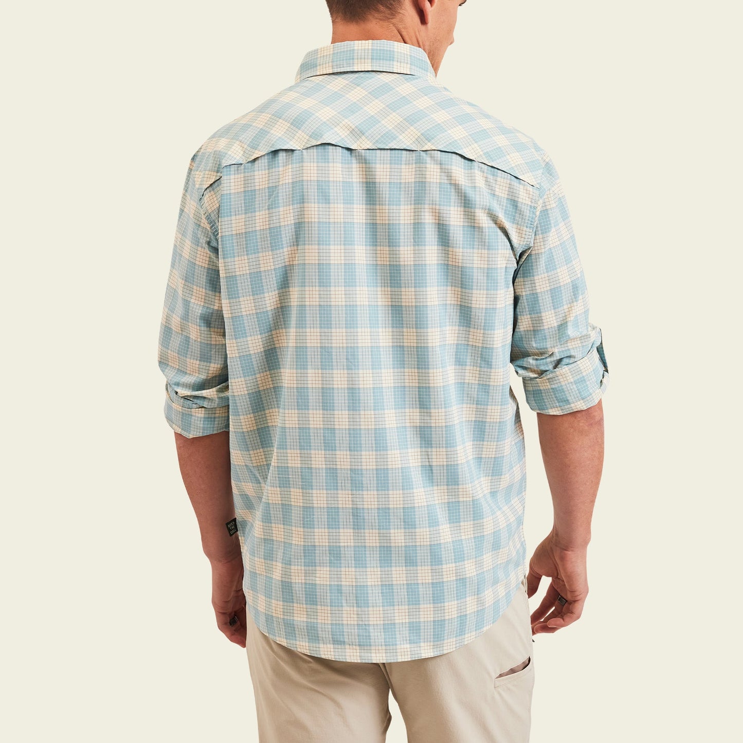 A man is shown from the back wearing a light blue and white plaid Matagorda Shirt with rolled-up sleeves and khaki pants, standing against a plain light background. This travel shirt is quick drying, perfect for any adventure.