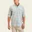A man wearing a light blue and beige plaid Matagorda Shirt with rolled-up sleeves and khaki pants, standing against a plain off-white background. This stylish travel shirt is quick drying, perfect for adventures on the go.
