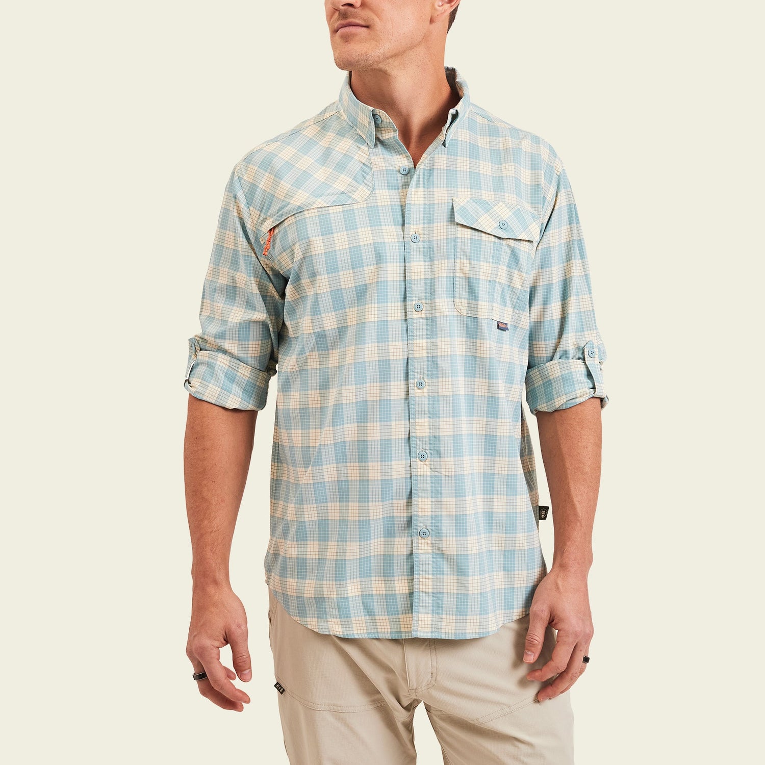 A man wearing a light blue and beige plaid Matagorda Shirt with rolled-up sleeves and khaki pants, standing against a plain off-white background. This stylish travel shirt is quick drying, perfect for adventures on the go.