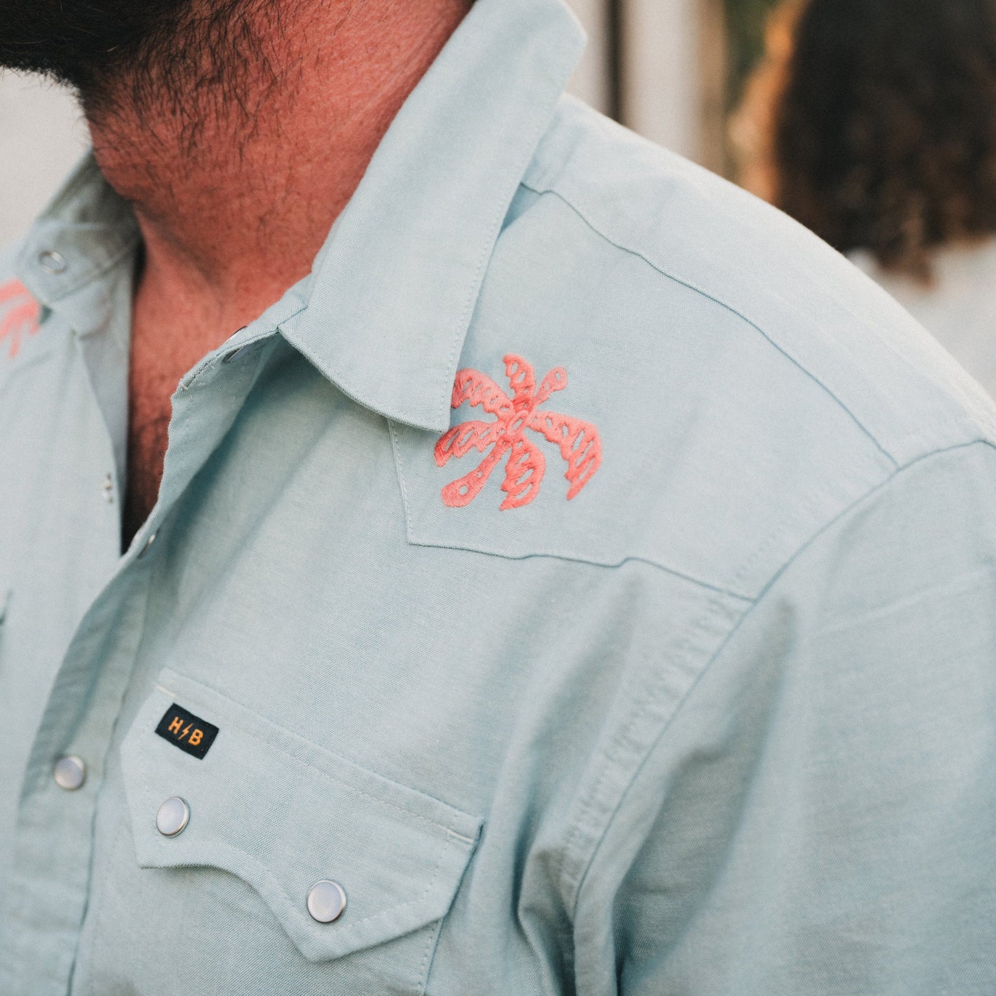 A close-up of a person wearing a light blue Crosscut Deluxe Shortsleeve summer shirt with pink palm tree motif embroidery on the shoulder and a small black HWB label above the pocket. The persons face is partially visible.
