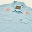 Light blue Crosscut Deluxe Shortsleeve western-style summer shirt with two front pockets, pearl snap buttons, and coral palm tree motif embroidery on the shoulders. A woven Howler brand label is visible inside the collar.