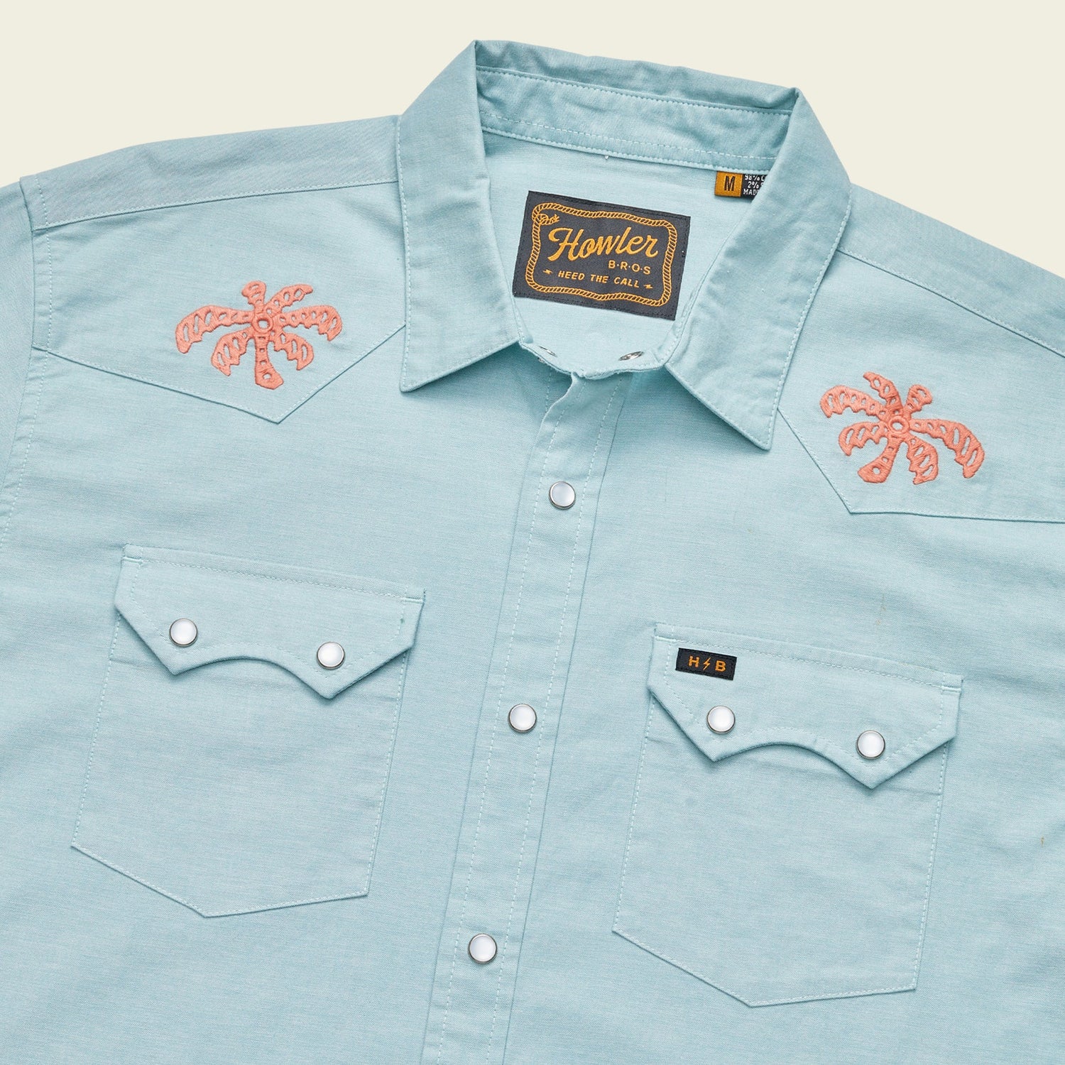 Light blue Crosscut Deluxe Shortsleeve western-style summer shirt with two front pockets, pearl snap buttons, and coral palm tree motif embroidery on the shoulders. A woven Howler brand label is visible inside the collar.