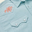 Close-up of a light blue Crosscut Deluxe Shortsleeve summer shirt with pointed collar and snap-button chest pocket, featuring an orange palm tree motif embroidery on the upper left side.