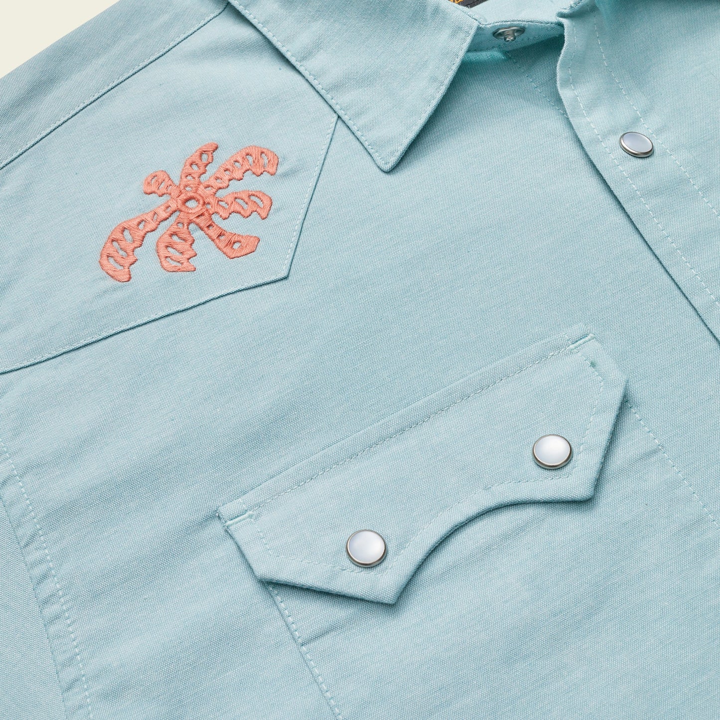 Close-up of a light blue Crosscut Deluxe Shortsleeve summer shirt with pointed collar and snap-button chest pocket, featuring an orange palm tree motif embroidery on the upper left side.