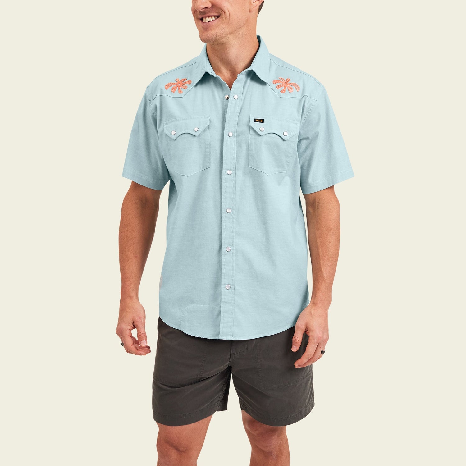 Smiling man wearing a light blue Crosscut Deluxe Shortsleeve summer shirt featuring orange lobster motif embroidery on the shoulders and dark gray shorts, standing against an off-white background.