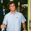 A man with curly brown hair in a light blue Crosscut Deluxe Shortsleeve stands in a bar holding a bottle of Corona beer, with neon signs and a fridge in the background.
