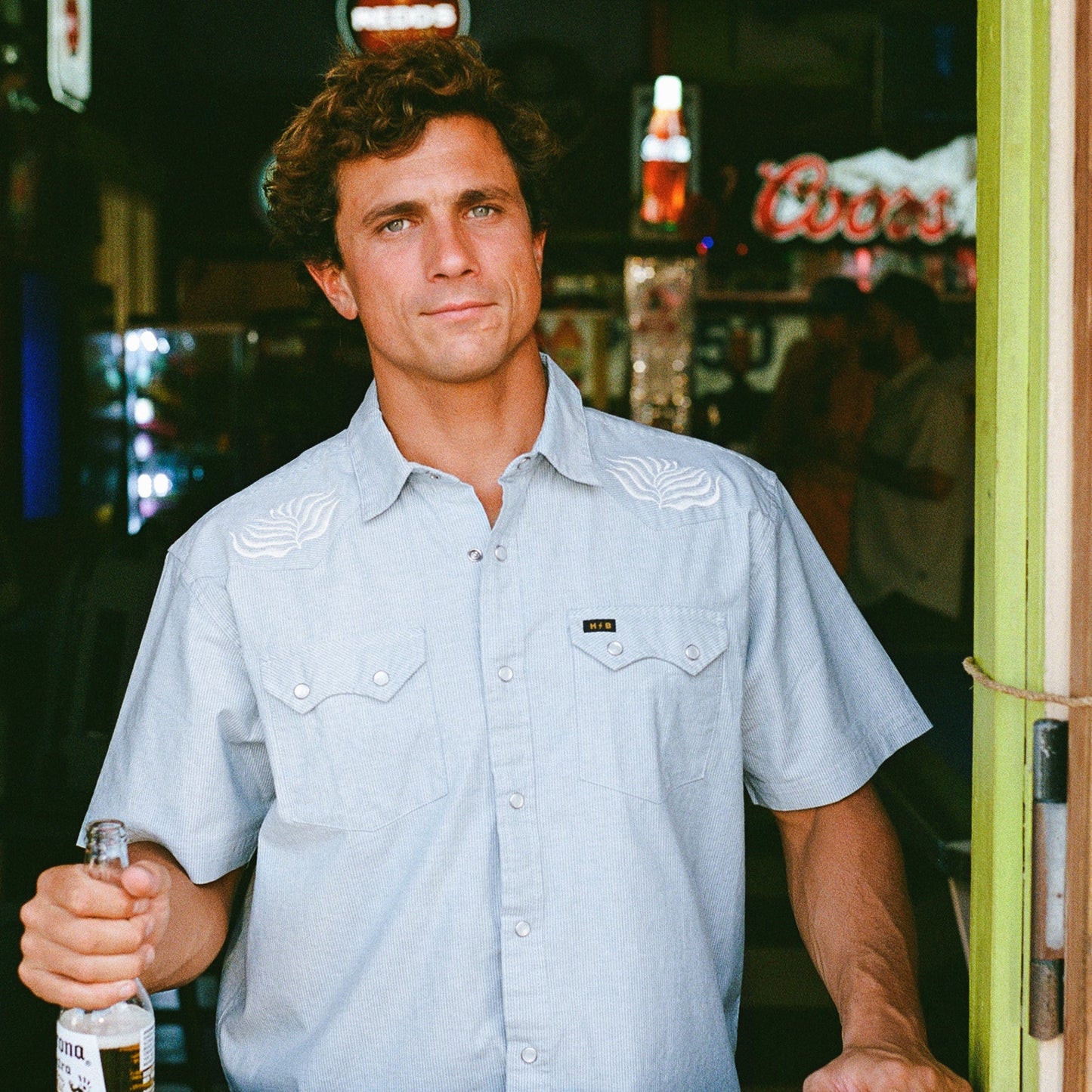 A man with curly brown hair in a light blue Crosscut Deluxe Shortsleeve stands in a bar holding a bottle of Corona beer, with neon signs and a fridge in the background.