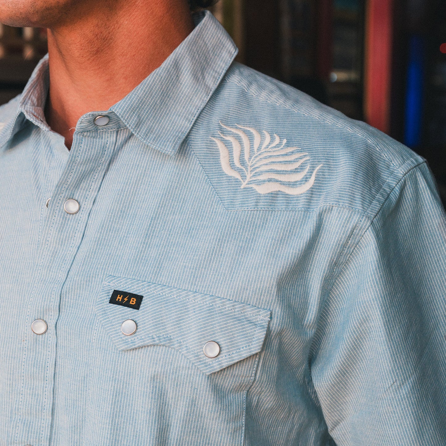 A close-up of a person wearing a light blue Crosscut Deluxe Shortsleeve with thin white stripes, motif embroidery on the shoulder, and a front pocket featuring a black and gold H+B tag.