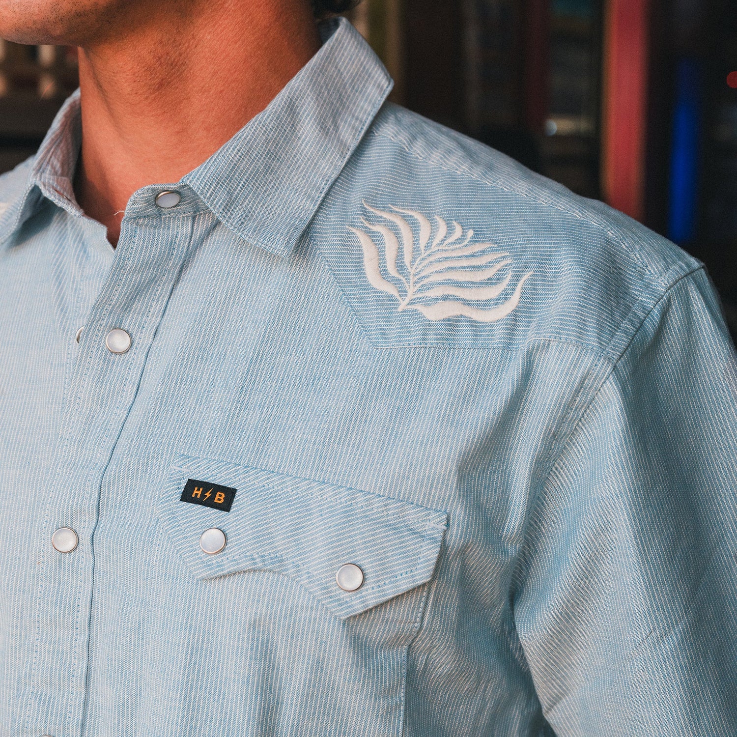 A close-up of a person wearing a light blue Crosscut Deluxe Shortsleeve with thin white stripes, motif embroidery on the shoulder, and a front pocket featuring a black and gold H+B tag.