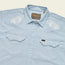 A light blue, striped Crosscut Deluxe Shortsleeve pearl snap shirt with pointed pockets and white motif embroidery on the shoulders. The shirt features a Howler brand tag on the inside collar.
